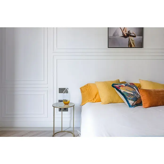 Nmc Decoflair Wandleiste SP2 - 2 m x 9 mm x 22 mm, Hochverdichteter Polyurethanschaum (PU) - Elegantes Schlafzimmer mit weißen Wandpaneelen, einem goldenen Beistelltisch mit Glasvase und farbenfrohen Kissen auf dem Bett.