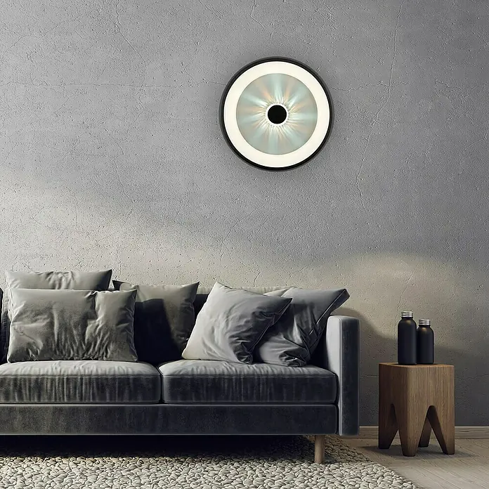 Just Light LED-Deckenleuchte rund VERTIGO - 33 W, Ø x H: 46,5 x 9,5 cm, Schwarz, Mehrfarbig - Wandleuchte mit kreisförmigem Design, bestehend aus einer schwarzen Umrandung und einem abstrakt gemusterten, beleuchteten Inneren, das über einem grauen Sofa hängt.