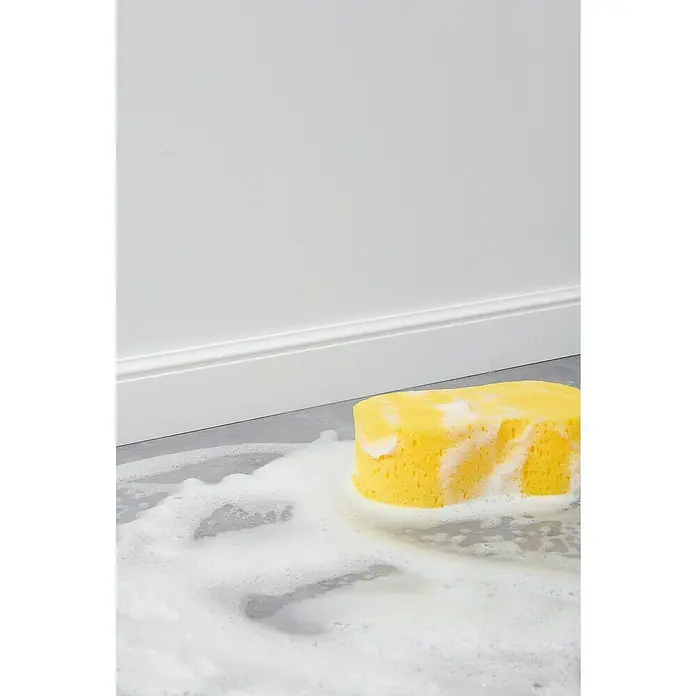 Nmc Decoflair Sockelleiste FL1 - 2 m x 13 mm x 80 mm - Gelber Reinigungsschwamm mit Seifenlauge auf grauem Boden vor einer weißen Wand. Perfekt für die Reinigung im Haushalt.