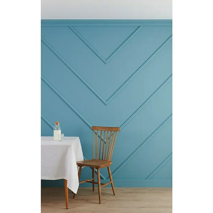 Nmc Decoflair Zierprofil Deckenleiste C - 2 m x 66 mm x 83 mm, Extrudiertes Polystyrol (XPS) - Eine Holzsessel steht neben einem Tisch mit einer weißen Tischdecke und einer Milchflasche vor einer blauen Wand mit Dekorleisten.