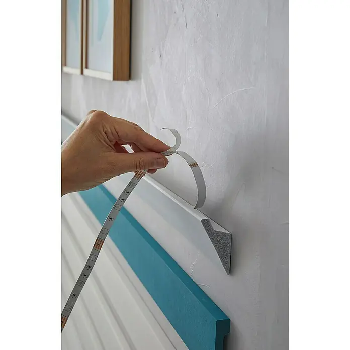 Nmc Decoflair Lichtleiste IL4 - 2 m x 36 mm x 60 mm, Hochverdichtetes extrudiertes Polymer (HDPS) - LED-Streifen wird auf einer Wandleiste angebracht, wobei die Schutzfolie abgezogen wird, um die Klebefläche freizulegen.