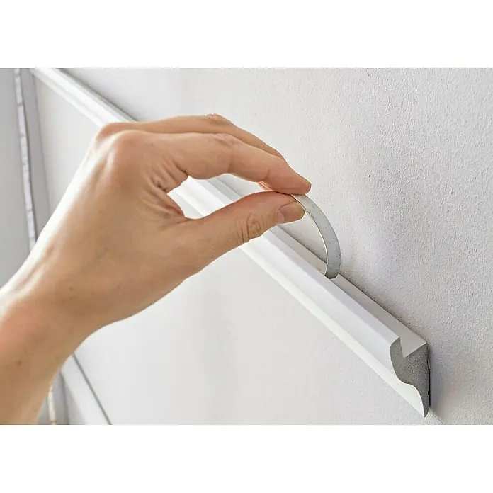Nmc Decoflair Lichtleiste IL3 - 2 m x 33 mm x 50 mm, Hochverdichtetes extrudiertes Polymer (HDPS) - Eine Hand befestigt eine weiße Zierleiste an einer Wand mit einem Klebestreifen, um das Produkt hervorzuheben.