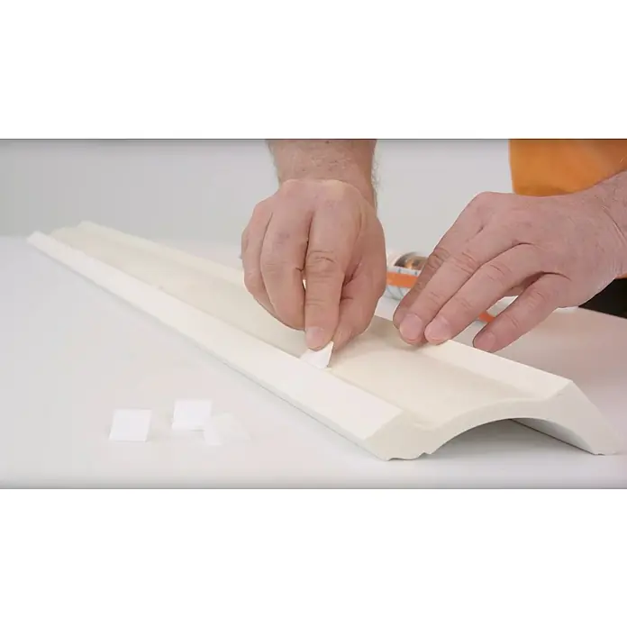 Nmc Decoflair Befestigungsclip für LED-Band - 20 Stk. - Eine Person befestigt Schaumstoffstücke an einer weißen Zierleiste. Die Leiste ist lang und hat ein geschwungenes Profil.