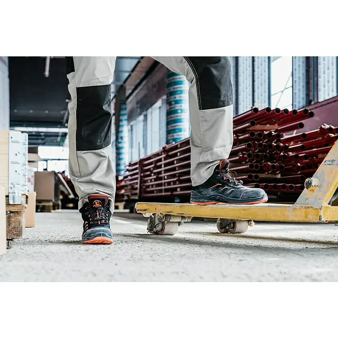 Botas de seguridad Gordon - 43, Clase de protección según EN ISO 20345: S1P, Negro/Naranja - Primer plano de botas de trabajo con protección de tobillo y detalles naranjas, usadas por una persona con pantalones de trabajo grises.