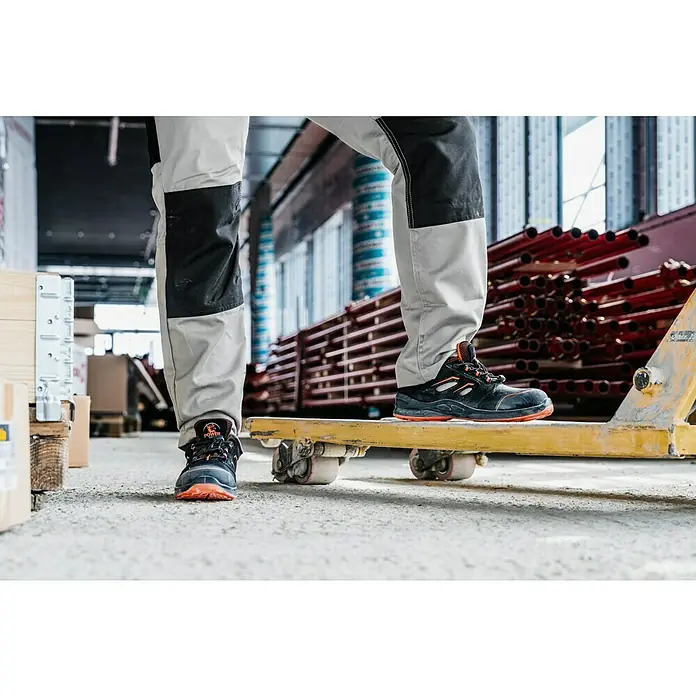 Sicherheitshalbschuh Tony - 42, Schutzklasse nach EN ISO 20345: S1P, Schwarz, Orange - Detailaufnahme von Arbeitsschuhen mit orangefarbenen Akzenten und strapazierfähiger Sohle, getragen auf einer gelben Plattform in industrieller Umgebung.