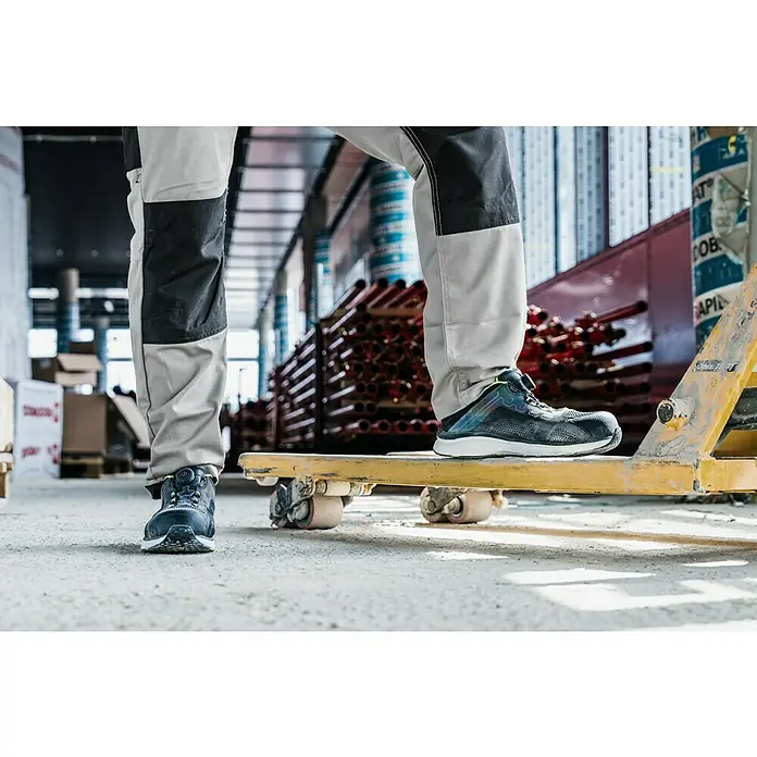 Zapato bajo de seguridad Fynn - Zapatos bajos, 43, Clase de protección según EN ISO 20345: S1P, Negro/Amarillo flúor - Zapatos de trabajo en foco: un usuario con calzado de seguridad está de pie en una plataforma amarilla en un almacén.