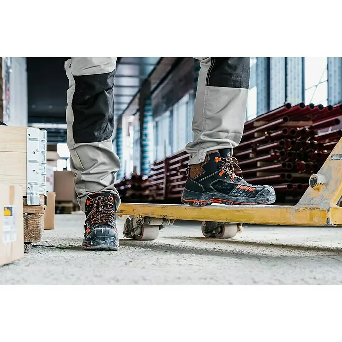 Botas de seguridad Maddox - 43, Clase de protección según EN ISO 20345: S3, Negro/Naranja - Primer plano de botas de trabajo sobre plataforma amarilla, ideales para la construcción. Botas robustas con detalles naranjas.