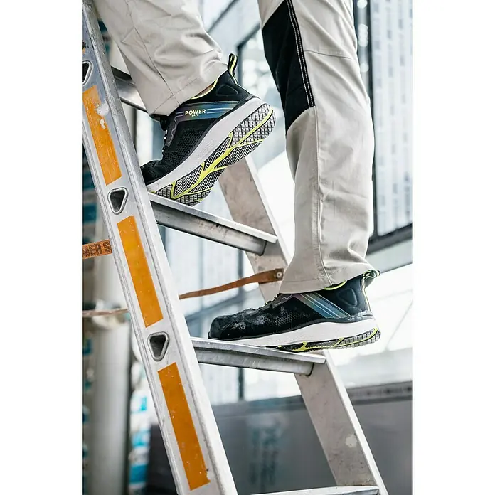Zapato bajo de seguridad Fynn - Zapatos bajos, 43, Clase de protección según EN ISO 20345: S1P, Negro/Amarillo flúor - Un hombre sube una escalera con zapatos de seguridad. Los zapatos son negros con detalles amarillos.