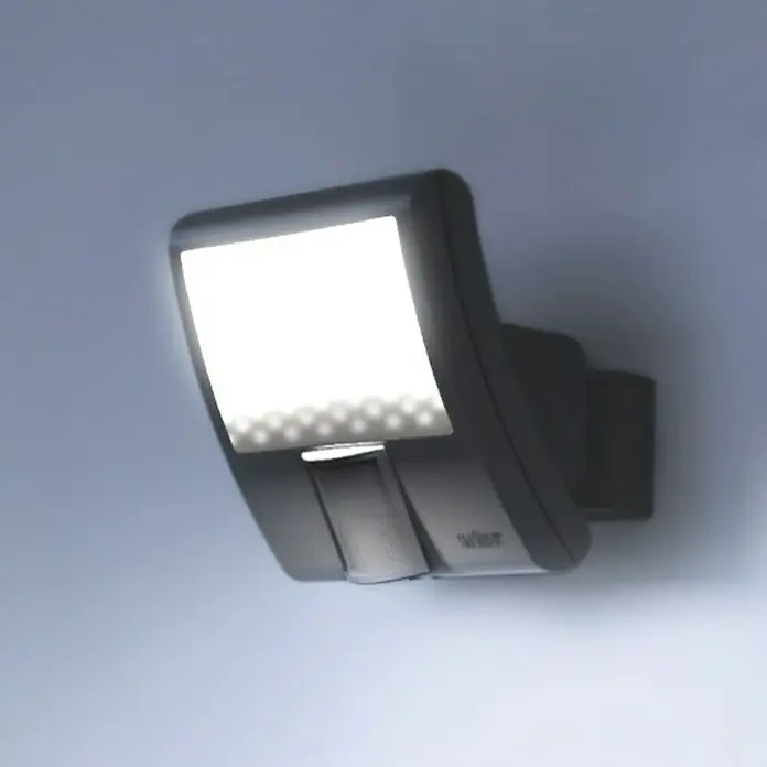 Steinel LED-Sensor-Strahler XLED home Curved S anthrazit - 9,3 W, L x B x H: 16 x 10,7 x 12 cm, Anthrazit, IP44 - Eine Nahaufnahme einer modernen, grauen Außenleuchte mit einem hell leuchtenden, rechteckigen Lichtfeld an einer Wand.