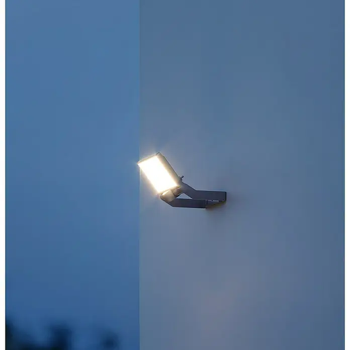 Steinel Proyector LED XLED ONE - 17,8 W, L x An x Al: 17,3 x 22,9 x 19,5 cm, Acero inoxidable, Blanco cálido - Una luz exterior moderna está montada en una pared, brillando intensamente. La luminaria es de color gris y rectangular.