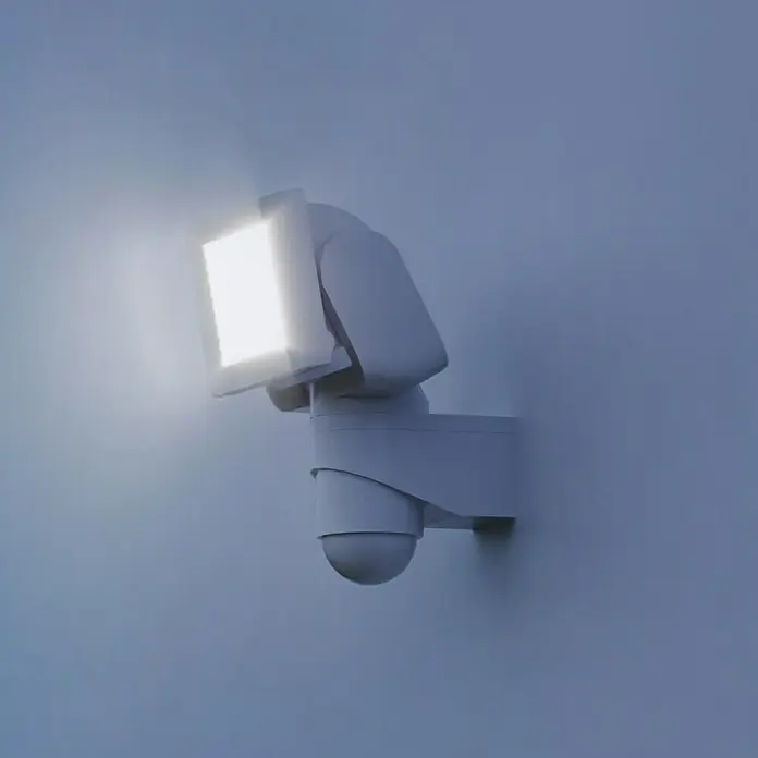 Steinel LED-Sensor-Strahler LS 150 S weiß - 14,7 W, L x B x H: 17 x 15,5 x 21,5 cm, Weiß, IP44 - Nahaufnahme einer weißen LED-Außenleuchte mit Bewegungsmelder an einer Wand, die helles Licht ausstrahlt.