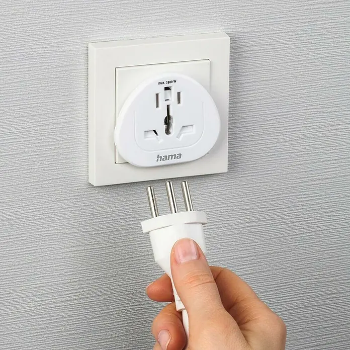Hama Reiseadapter Welt nach Europa - Netzsteckertyp Reiseadapter: Typ E+F (CEE 7/7), Weiß - Nahaufnahme eines weißen Netzadapters von Hama mit einem Stecker, der in eine Steckdose gesteckt wird, vor einer grauen Wand.