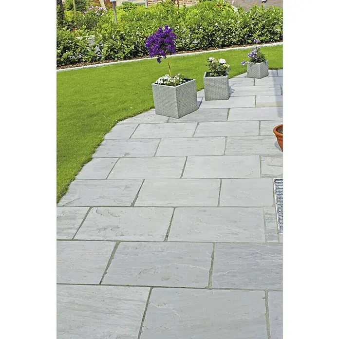 Terrassenplatte Delhi Grey - 60 x 30 x 2,5 cm, Grau, Sandstein - Ein gepflasterter Weg führt durch einen Garten, flankiert von Blumenkübeln, die mit bunten Blumen bepflanzt sind.