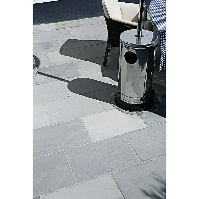 Terrassenplatte Delhi Grey - 60 x 30 x 2,5 cm, Grau, Sandstein - Eine moderne Terrassenheizung aus Edelstahl steht auf einem gepflasterten Bereich neben einem Stuhl und sorgt für Wärme im Freien.