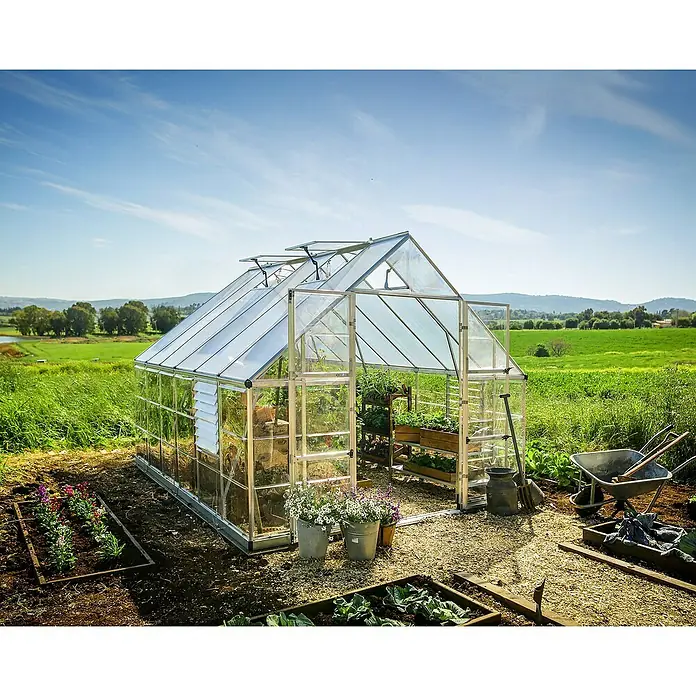 Palram – Canopia Invernadero Balance - Medidas ext. (An x Pr): 304 x 366 cm, 0,7 mm, Plateado - Primer plano de un invernadero con plantas y herramientas de jardinería al aire libre, rodeado de naturaleza verde y cielo azul.