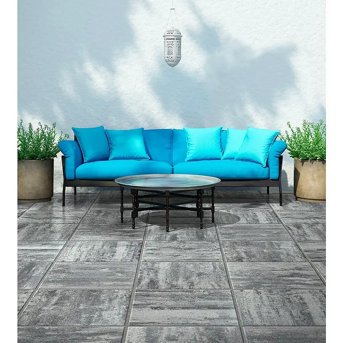 Terrassenplatte Saturn - 50 x 50 x 4 cm, Grau/Schwarz, Beton - Outdoor-Szene mit einem blauen Sofa, einem runden Tisch, Pflanzen in Töpfen und einer dekorativen Laterne vor einer weißen Wand.