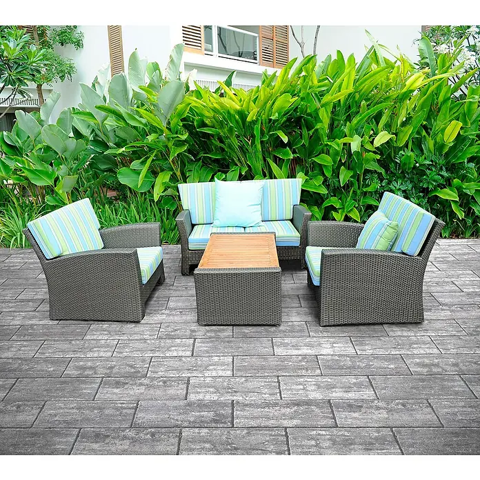 Terrassenplatte Latina - 48 x 24 x 3 cm, Grau/Schwarz, Beton - Dieses Bild zeigt ein graues Rattansofa-Set mit gestreiften hellblauen und hellgrünen Kissen auf einer Terrasse vor üppigem Grün.