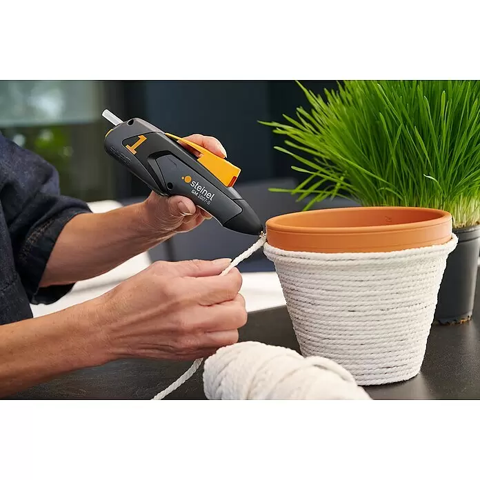 Steinel Akku-Heißklebestift MobileGlue 1007-S - 3,6 V, Durchmesser Klebepatrone: 7 mm, 170 °C - Eine Person verwendet eine grau-orange Steinel Heißklebepistole, um weißes Seil an einen Terrakotta-Topf zu kleben.