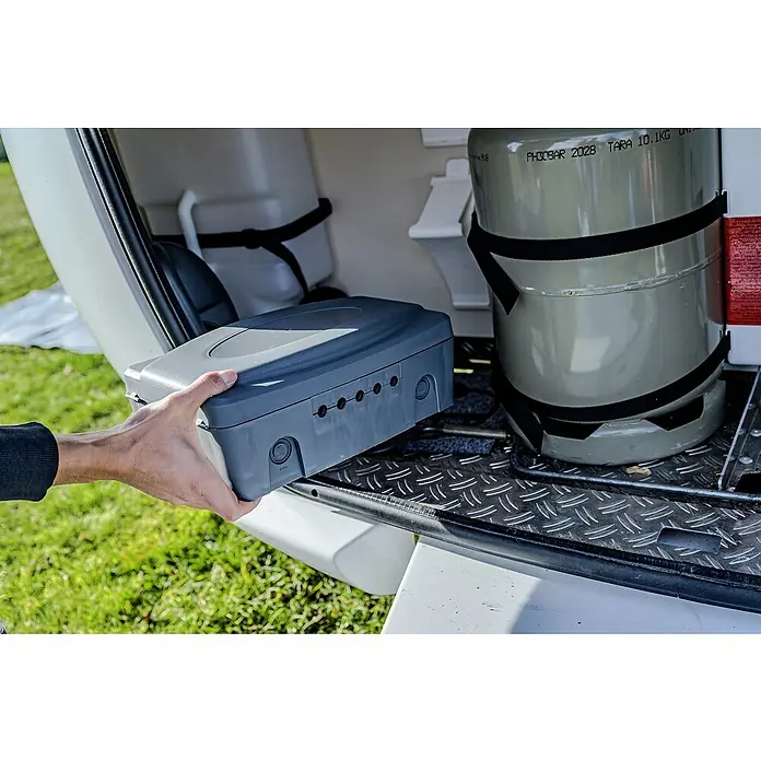 Brennenstuhl Kabelbox - L x B x H: 34 x 23 x 12,5 cm, IP54 - Die Hand hält eine graue Kiste mit Löchern in einem Wohnmobil, daneben eine Gasflasche. Perfekt für Campingausflüge.