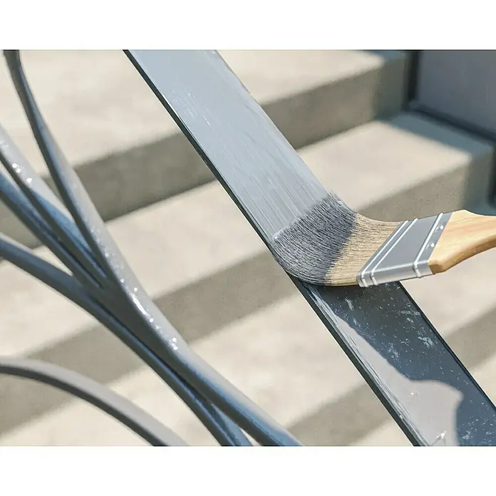 swingcolor Buntlack Kunstharz für Außen - Silbergrau, Hochglänzend, 750 ml, Lösemittelbasiert - Nahaufnahme eines Holzgriffpinsels, der auf einem Metallgeländer graue Farbe aufträgt, mit Betonstufen im Hintergrund.