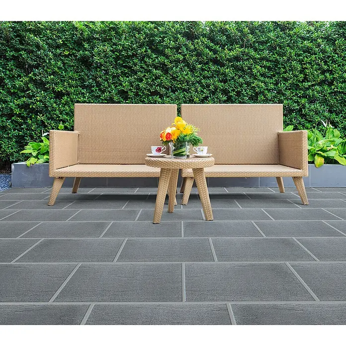 Terrassenplatte Wild Stone - 60 x 40 x 4 cm, Titanio, Beton - Dieses Bild zeigt eine stilvolle Rattan-Garnitur mit Tisch und Blumen. Ideal für den Garten oder die Terrasse, lädt sie zum Verweilen ein.