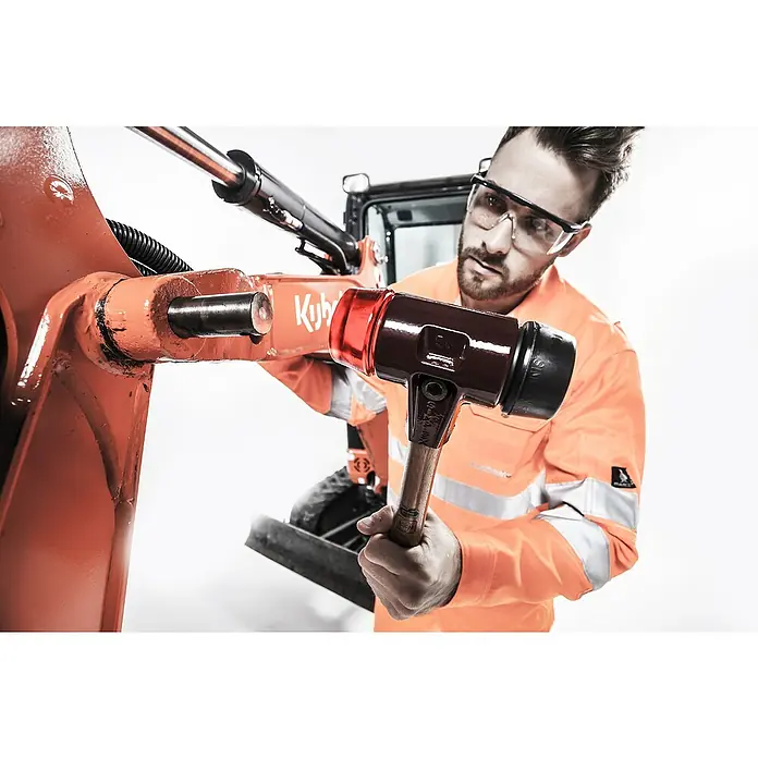Halder Simplex Schonhammer GUKO - Durchmesser Kopf: 50 mm - Ein Mann verwendet einen Schonhammer, um einen Bolzen an einem orangefarbenen Kubota-Bagger zu montieren. Schutzkleidung ist ein Muss.