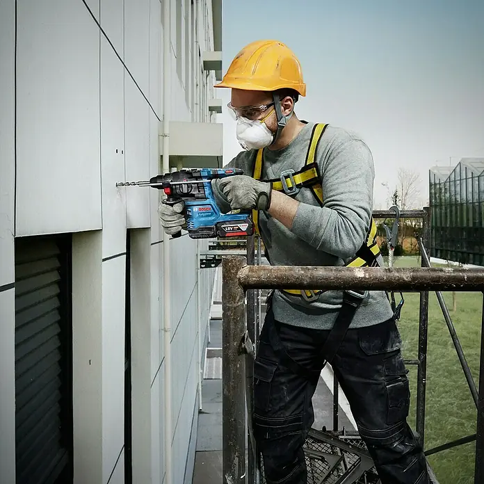 Bosch Professional AMPShare 18V Akku-Bohrhammer GBH 18V-22 - 18 V, Ohne Akku, 1,9 J - Ein Bauarbeiter bohrt mit einem blauen Bosch-Bohrhammer ein Loch in eine Wand. Er trägt Schutzkleidung wie Helm und Maske.