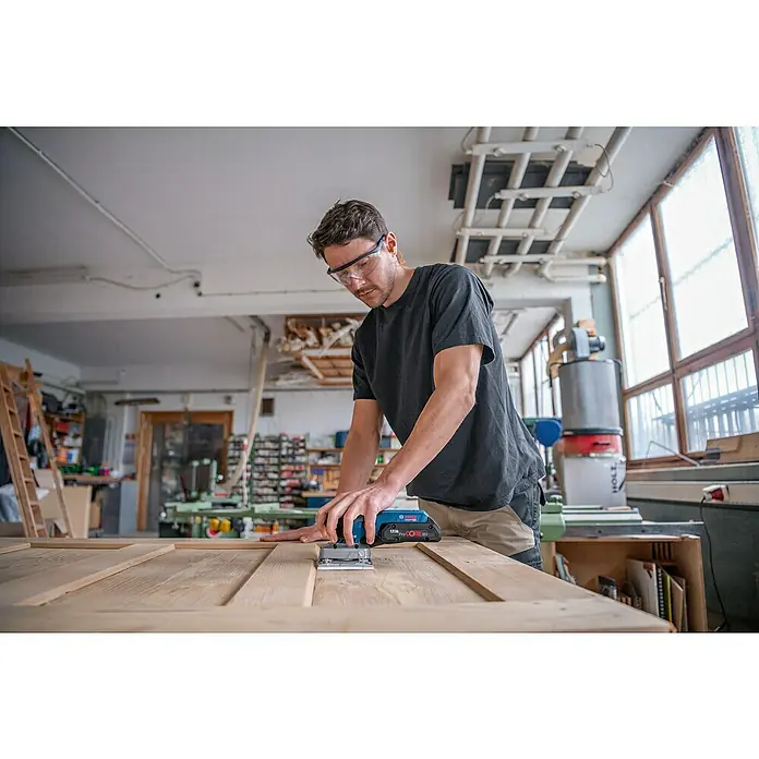 Bosch Professional AMPShare 18V Akku-Schwingschleifer GSS 18V-13 - 18 V, Ohne Akku, Leerlaufschwingzahl: 12.000 U/min - 20.000 U/min - Ein Mann, der einen Bosch Akku-Multischleifer benutzt, um eine Holzoberfläche in einer Werkstatt zu schleifen. Er trägt eine Schutzbrille.
