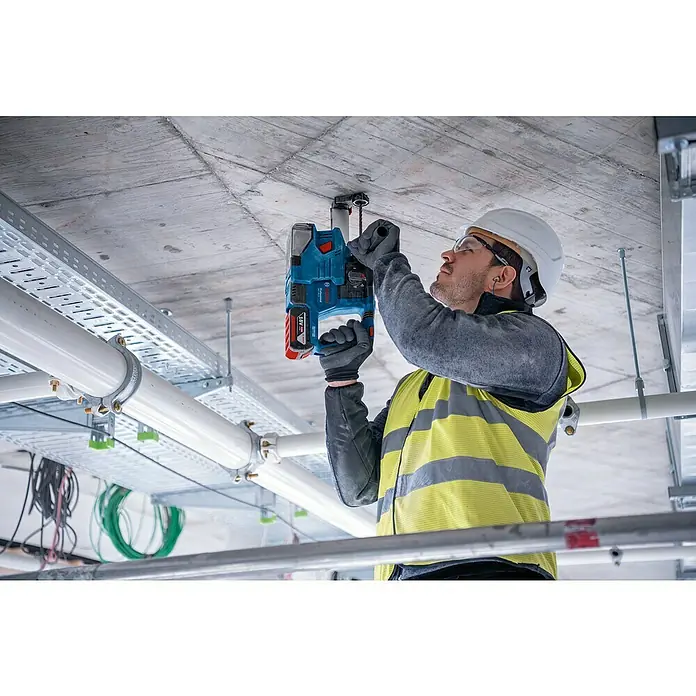 Bosch Professional Staubabsaugung GDE 18V-12 - Passend für: Bosch Professional Bohrhammer GBH 18V-22 - Ein Bauarbeiter befestigt mit einer blauen Bosch-Akku-Bohrhammer ein Element an einer Betondecke, umgeben von Rohren und Kabelkanälen.