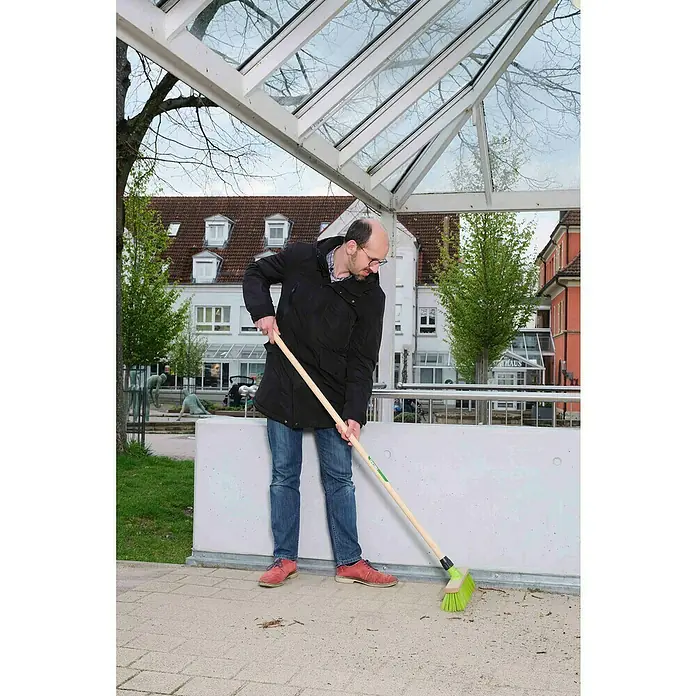 Freund Victoria Besen - 40 cm, Material Borsten: Haarmischung - Ein Mann mit Brille kehrt mit einem grünen Besen eine Fläche im Freien. Der Holzstiel und die kräftigen Borsten helfen beim Säubern.