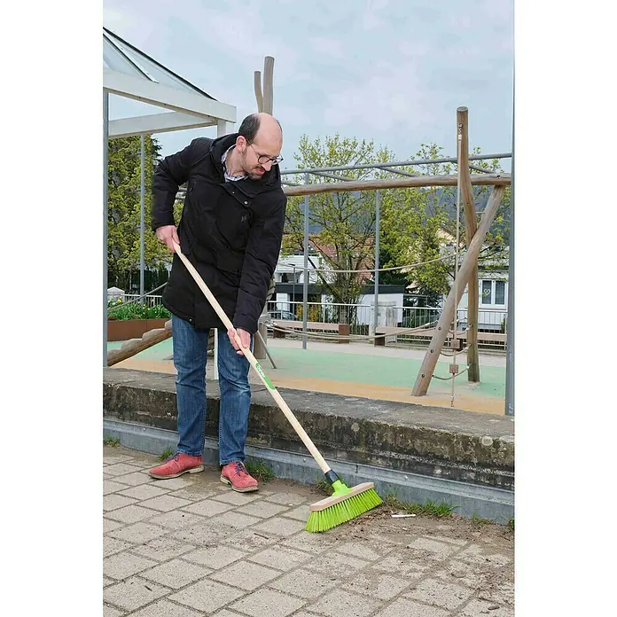 Freund Victoria Besen - 40 cm, Material Borsten: Haarmischung - Mann kehrt einen Bereich mit einer hellgrünen Straßenbürste mit einem Holzstiel auf einer gepflasterten Fläche im Freien.