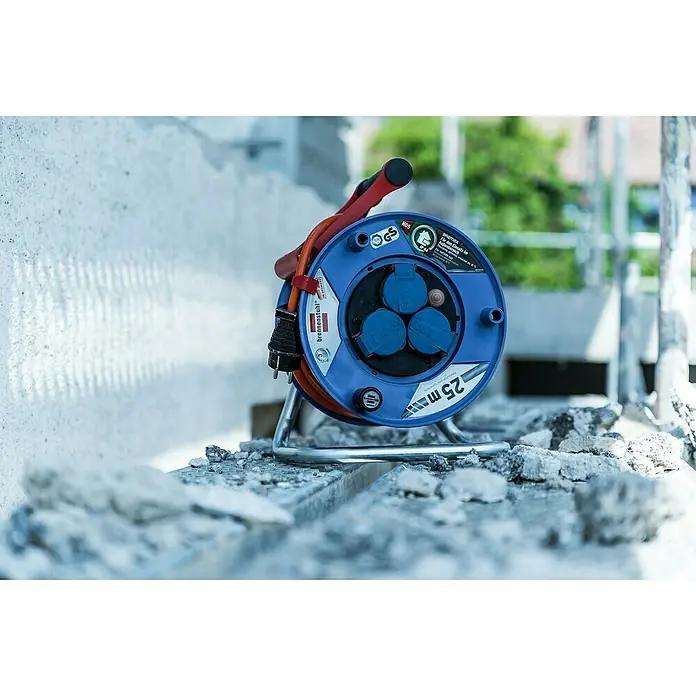 Brennenstuhl Garant Kabeltrommel - Kunststoff, 25 m, AT-N05V3V3-F, IP44 - Blaue Brennenstuhl Kabeltrommel mit rotem Kabel und 3 blauen Steckdosen, auf einer Baustelle mit Steinen im Vordergrund.