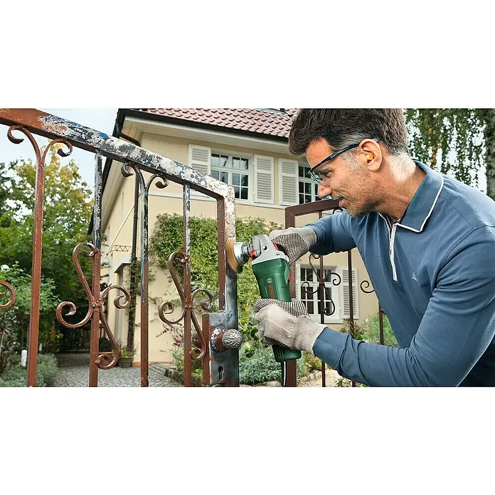 Bosch Universal Amoladora angular eléctrica Grind 750-115 - 750 W, Diámetro disco: 115 mm, 12.000 r.p.m. - Un hombre elimina el óxido de una valla metálica frente a una casa, utilizando una amoladora de cepillo de alambre Bosch verde.