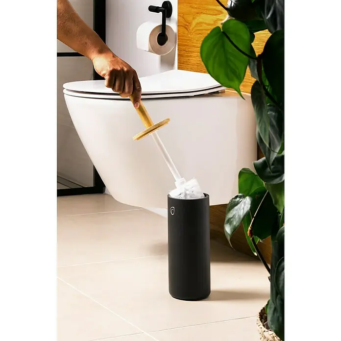 Camargue WC-Bürstengarnitur Vero - Bambus, Anthrazit, Höhe: 38 cm - Eine Hand legt eine Toilettenbürste mit Holzgriff zurück in den schwarzen Halter neben einer Toilette und einer Pflanze.