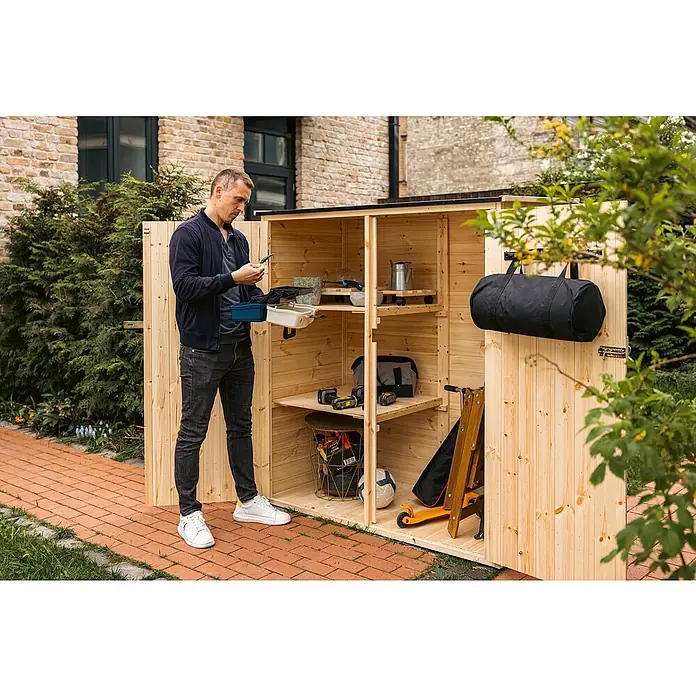 Upyard Geräteschrank Classic Line - 150 x 80 cm, Holz, Natur - Mann vor einem Holzgartenhaus mit Regalen, Werkzeug und Sportartikeln. Das Gartenhaus bietet Stauraum für Gartengeräte und mehr.