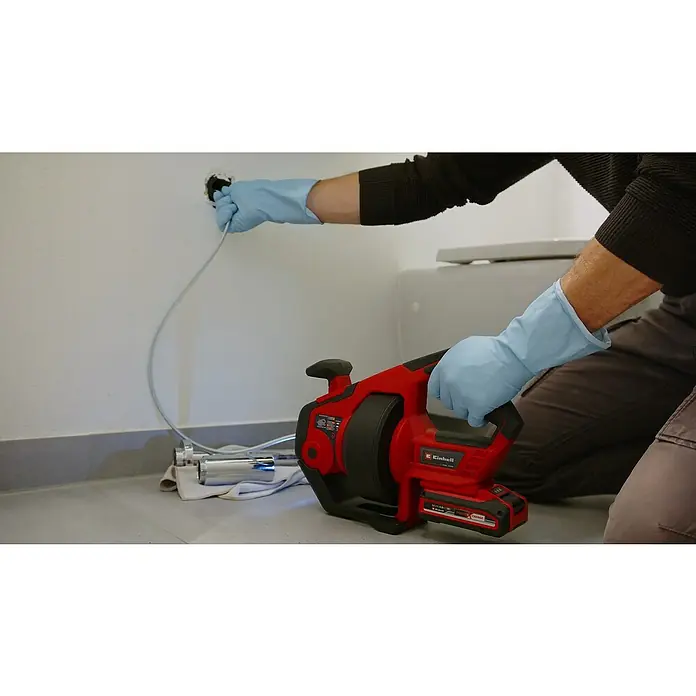 Einhell PXC 18V Desatascador de batería TE-DA 18/760 Li Solo - 18 V, Sin batería - Un hombre usa un barreno de limpieza de desagües Einhell para despejar una tubería obstruida, con una herramienta roja inalámbrica y guantes azules.