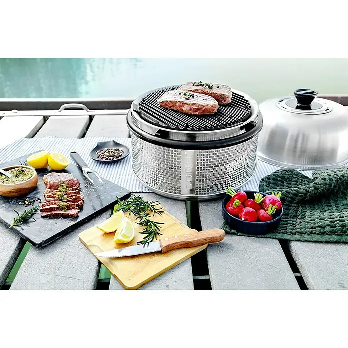 Cobb Holzkohlegrill Air Deluxe - Durchmesser Grillfläche: 32 cm, Silber - Bild eines Cobb-Grills mit Steaks, Radieschen und Zitrone auf einem Tisch mit Meerblick.