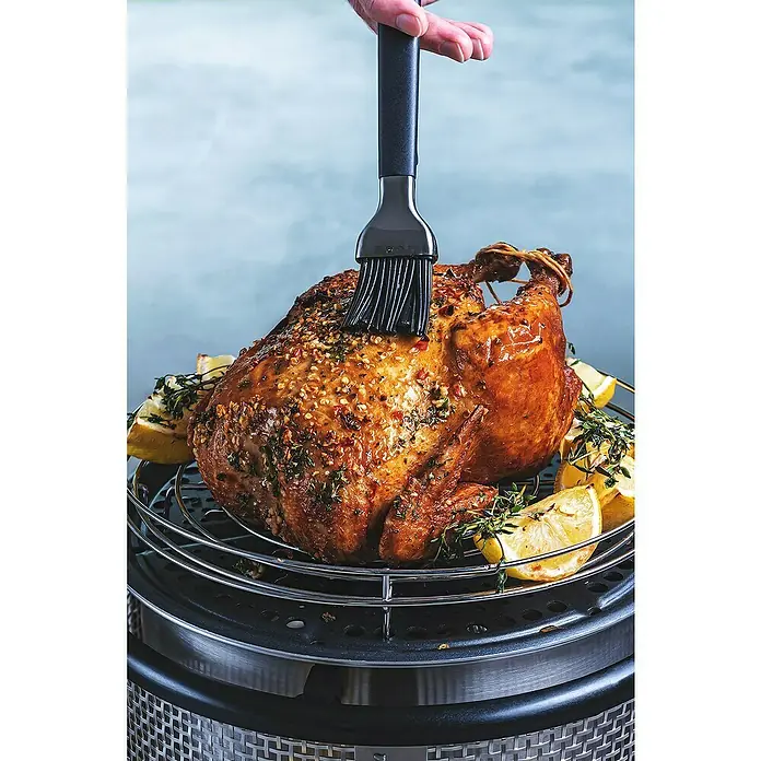 Cobb Grillrost - Durchmesser: 28,5 cm - Ein saftiges Brathähnchen wird mit einer schwarzen Bürste beträufelt und liegt auf einem Rost mit Zitronenspalten.
