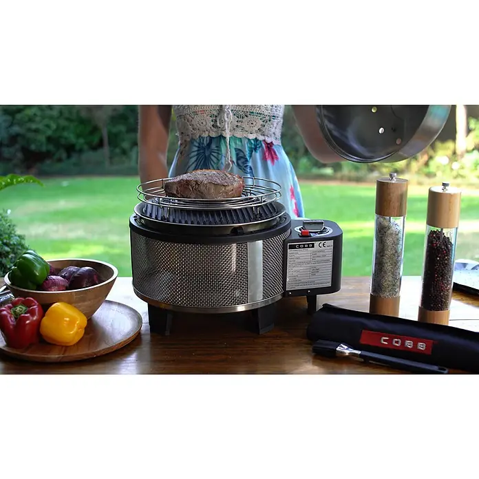Cobb Gasgrill Deluxe - Durchmesser Grillfläche: 32 cm, 1,23 kW, Anzahl Brenner: 1 - Ein Cobb-Grill mit Steak, Gemüse, Gewürzmühlen und Zubehör auf einem Tisch im Freien.