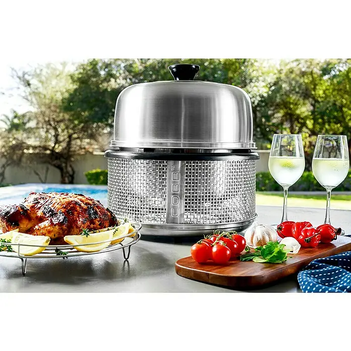 Cobb Holzkohlegrill Air DeluxeDurchmesser Grillfläche: 32 cm, Silber - Ein Cobb Grill mit leckerem Hähnchen, Tomaten und Wein auf einem Tisch im Freien. Ideal für ein entspanntes Barbecue im Garten.