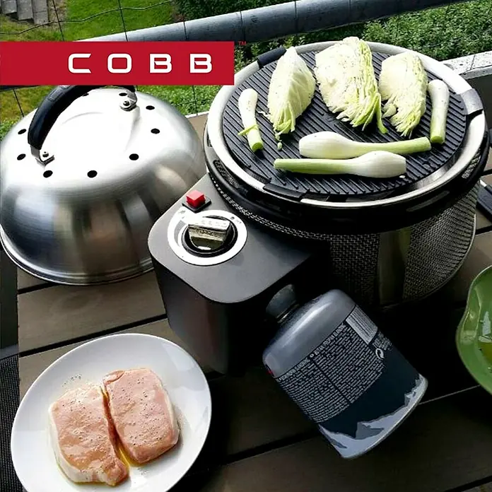 Cobb Gasgrill Deluxe - Durchmesser Grillfläche: 32 cm, 1,23 kW, Anzahl Brenner: 1 - Der Cobb-Grill, bereit zum Grillen von Kohl und Frühlingszwiebeln, neben einer Platte mit Fleisch.