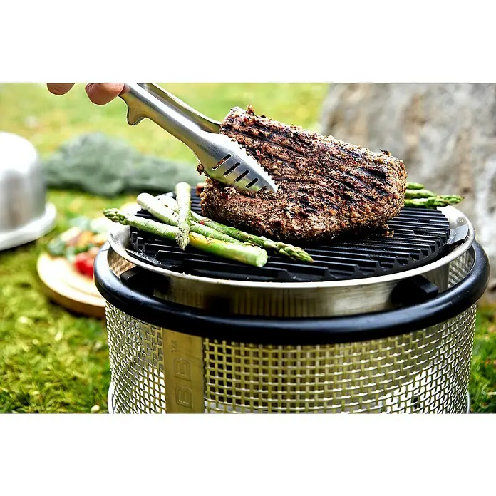 Cobb Holzkohlegrill Air Deluxe - Durchmesser Grillfläche: 32 cm, Silber - Ein saftiges Steak und grüner Spargel werden auf einem Grill mit Grillzange im Freien zubereitet.