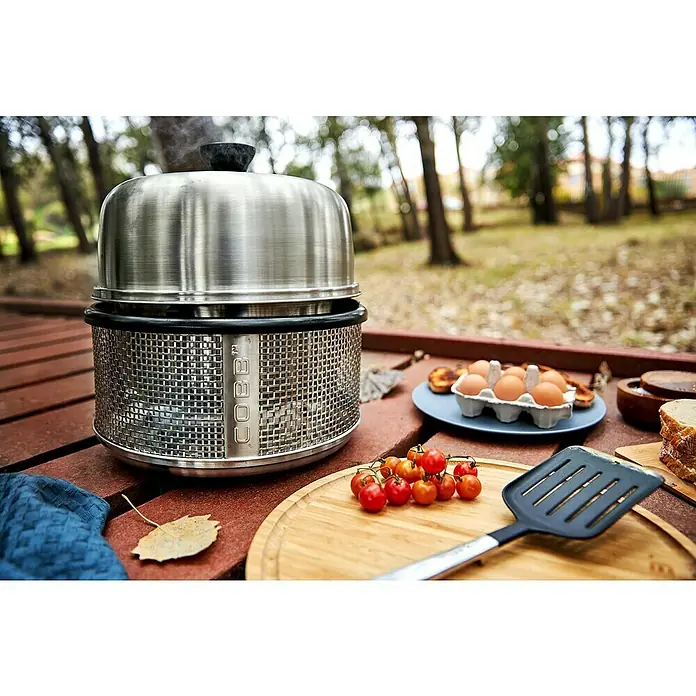 Cobb Holzkohlegrill Air Deluxe - Durchmesser Grillfläche: 32 cm, Silber - Ein Cobb Grill steht auf einem Tisch im Freien, umgeben von Zutaten wie Eiern und Tomaten für ein gemütliches Outdoor-Essen.