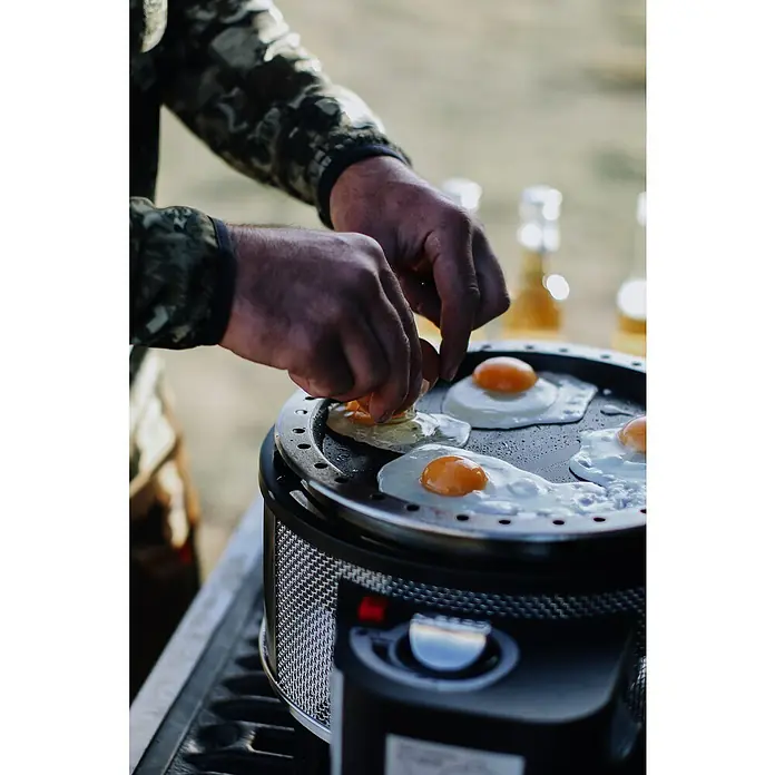 Cobb Gasgrill Deluxe - Durchmesser Grillfläche: 32 cm, 1,23 kW, Anzahl Brenner: 1 - Ein Mann bereitet auf einem tragbaren Kochfeld Spiegeleier zu. Das Kochfeld ist rund und hat eine schwarze Oberfläche.
