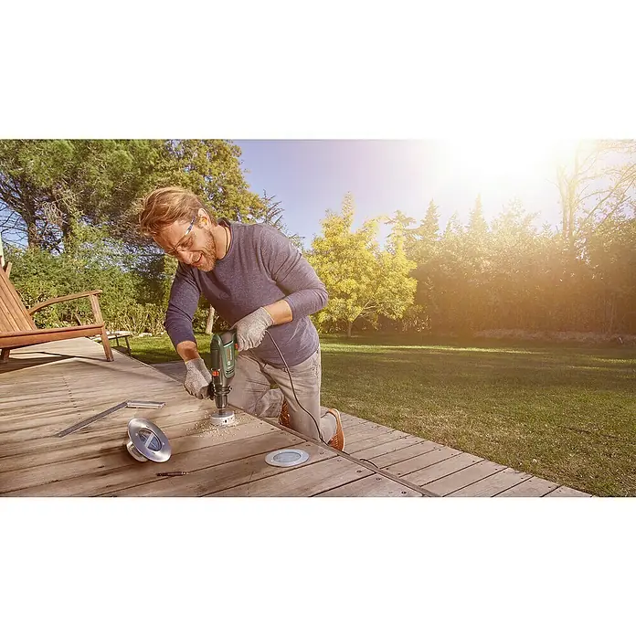 Bosch Universal Schlagbohrmaschine UniversalImpact 730730 W, Max. Schlagzahl: 48.000 /min - Ein Mann bohrt mit einer Bosch-Bohrmaschine ein Loch in ein Holzdeck, um eine Bodenleuchte zu installieren. Er trägt Schutzbrille und Handschuhe.