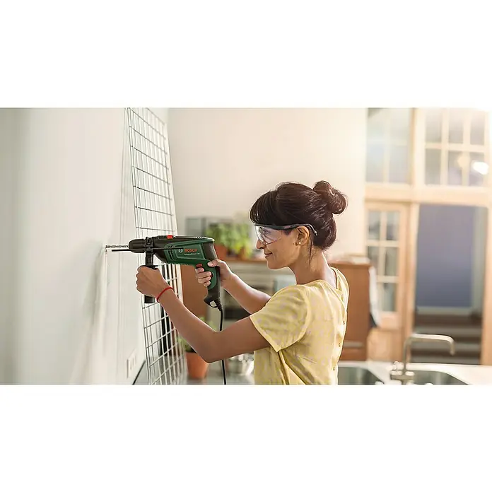 Bosch Universal Schlagbohrmaschine UniversalImpact 730 - 730 W, Max. Schlagzahl: 48.000 /min - Frau bohrt mit einer Bosch UniversalImpact 730 Bohrmaschine ein Loch in eine weiße Wand, während sie eine Schutzbrille trägt.
