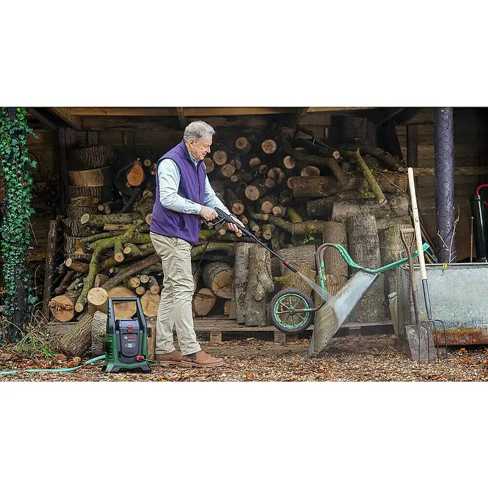 Bosch Power for All 36V Akku-Hochdruckreiniger UniversalAquatak 36V-100 - 5 -tlg., 36 V, Ohne Akku - Mann reinigt eine Schubkarre mit einem Bosch Hochdruckreiniger vor einem Stapel Holz.