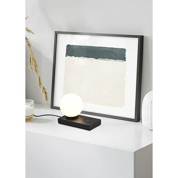 Reality Leuchten Tischleuchte Chloe - 40 W, L x B x H: 10 x 20 x 15 cm, Schwarz-matt, E14 - Elegante Szene mit einer modernen Kugel Lampe mit kabellosem Ladegerät und einem gerahmten abstraktem Kunstwerk auf einer weißen Oberfläche.
