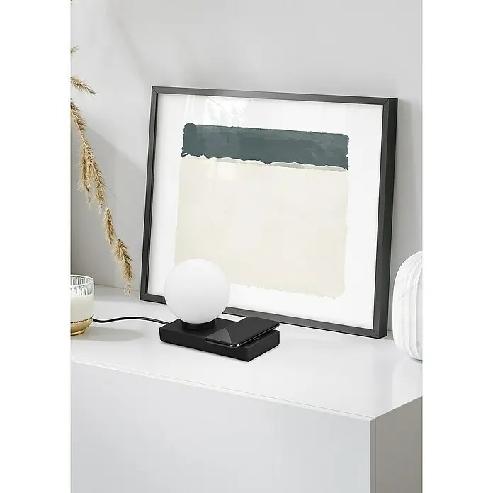 Reality Leuchten Tischleuchte Chloe - 40 W, L x B x H: 10 x 20 x 15 cm, Schwarz-matt, E14 - Nahaufnahme einer schwarzen Nachttischlampe mit integriertem kabellosem Ladegerät, platziert auf einem weißen Schrank vor einem gerahmten Kunstwerk.