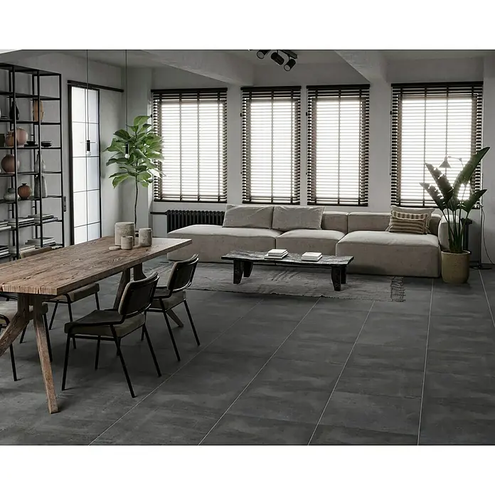 Feinsteinzeugfliese Manhattan Dark - 60 x 60 cm, Anthrazit, Matt - Ein Innenraum mit einem Holztisch und Stühlen im Vordergrund, einer Couch und Fenster mit Jalousien im Hintergrund.